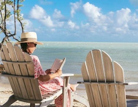 A woman in a sun hat sits on a beach chair reading a book by the shore, with two more chairs, sandy ground, and a bright blue sky.