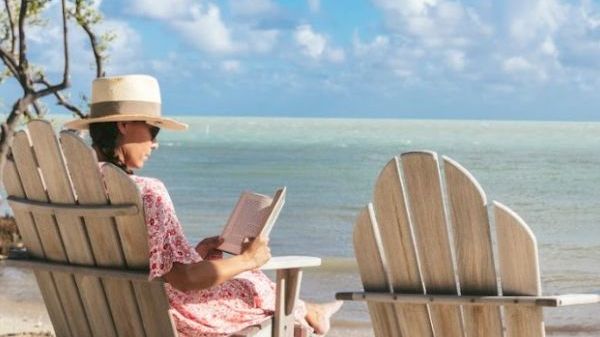 A person relaxes in an adirondack chair on the beach, reading a book under a sunny sky with gentle waves lapping at the shore.