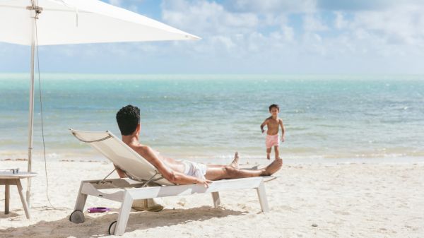 A man relaxes on a beach chair while a child joyfully runs towards the water under a bright blue sky. It's a perfect beach day.