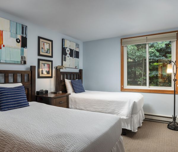 The image shows a cozy bedroom with two beds, abstract art, a window, and a warm wooden decor. It feels inviting and peaceful.