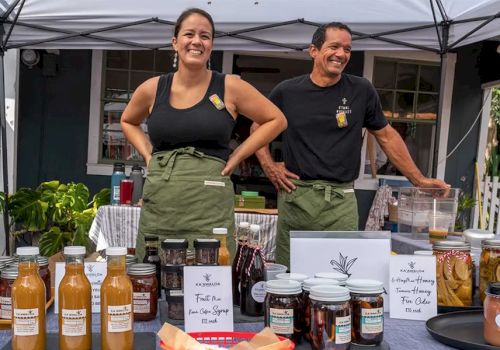 Two smiling vendors stand behind a table of bottled sauces, jams, and jars under a tent, with signs and samples on display.