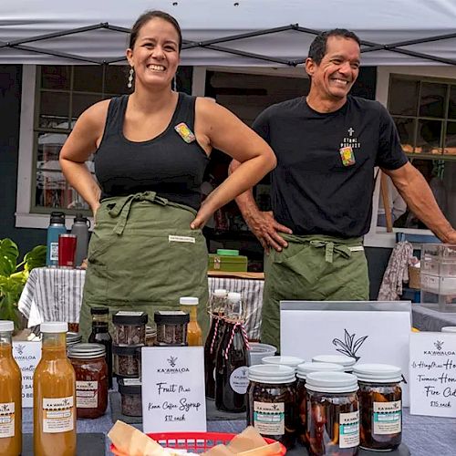Two smiling vendors stand behind a table of bottled sauces, jams, and jars under a tent, with signs and samples on display.