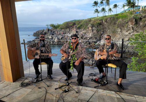 Three musicians perform outdoors near the ocean, wearing Hawaiian shirts and playing guitars, surrounded by tropical scenery.