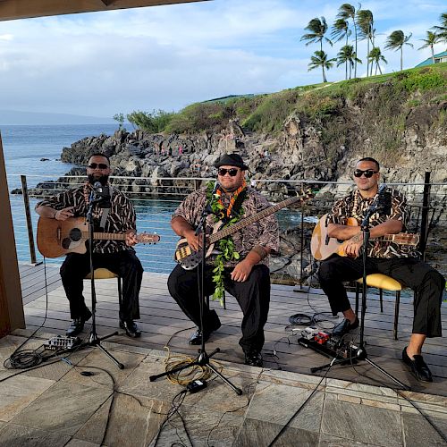 Three musicians perform outdoors near the ocean, wearing Hawaiian shirts and playing guitars, surrounded by tropical scenery.