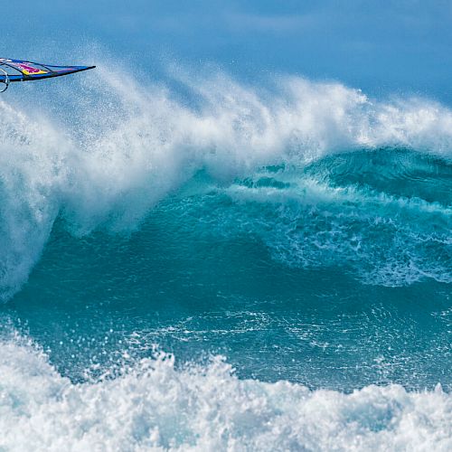 A windsurfer performs an acrobatic jump over vibrant blue waves, captured in a moment of thrilling action. The ocean sparkles.