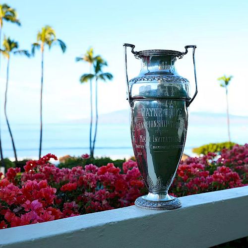 The image features a trophy resting on a ledge, surrounded by vibrant pink flowers, with a scenic ocean view and palm trees.