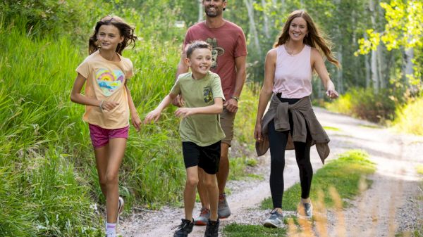 A family of four enjoys a walk along a scenic trail, surrounded by lush greenery and sunlight filtering through the trees.