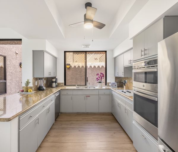 The image shows a modern kitchen with gray cabinetry, stainless steel appliances, a ceiling fan, and a view of an outdoor patio area.