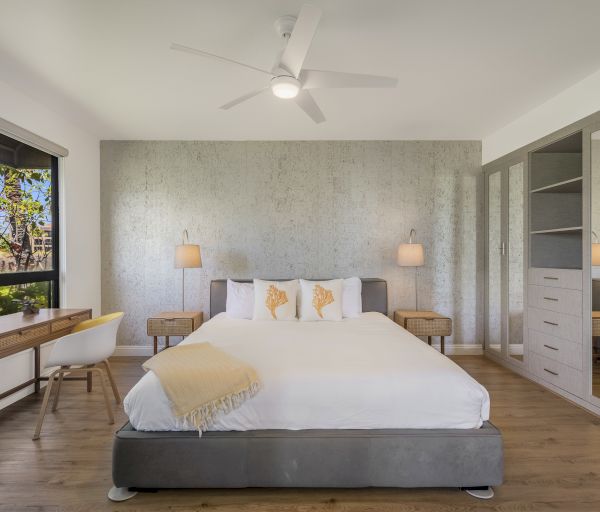 The image shows a modern bedroom with a large bed, stylish furniture, a desk, and bright natural light from a window.