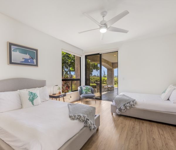 The image shows a bright room with two beds, a ceiling fan, and large windows overlooking a green landscape. It feels inviting and airy.