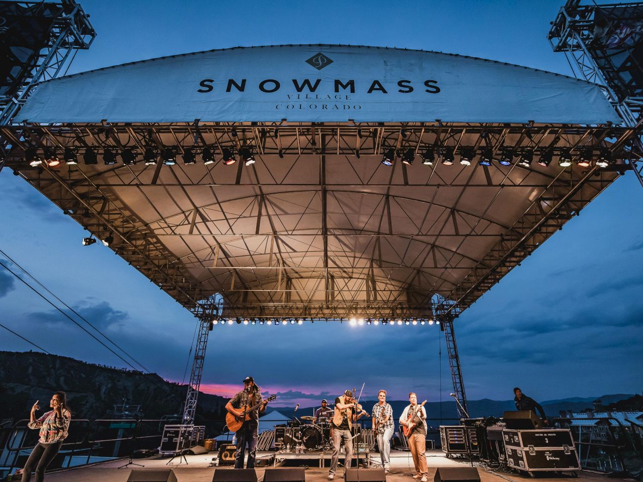 A band performs on stage at Snowmass Village, Colorado, under a vibrant sky as night approaches, creating a lively atmosphere.
