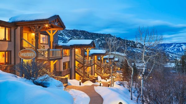 The image shows a cozy mountain lodge in winter, surrounded by snow, with warm lights glowing from the balconies and trees nearby.