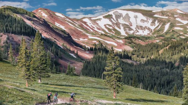 Three mountain bikers ride along a winding trail in a lush, green landscape surrounded by mountains and patches of snow.