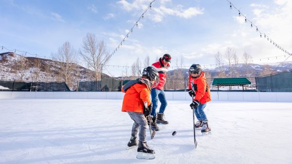 A father teaches two children to skate and play hockey on an outdoor rink, surrounded by snow and mountains under a bright sky.