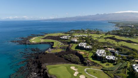An aerial view of a coastal golf course with lush greenery, rocky shoreline, and a clear blue ocean under a bright sky.