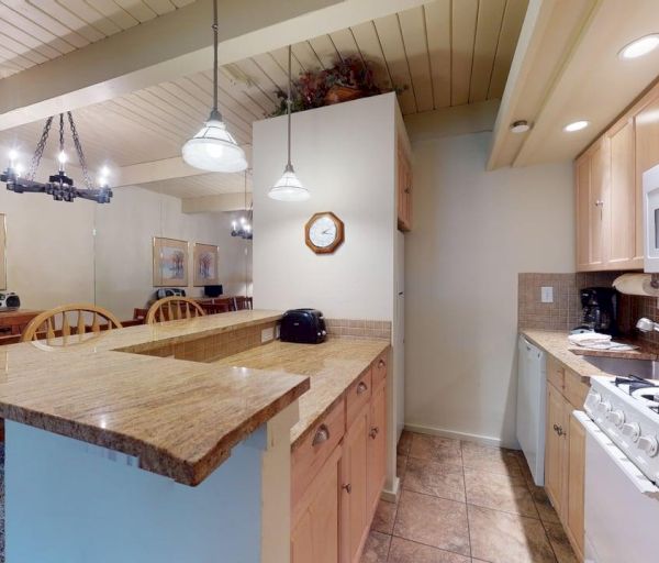 The image shows a modern kitchen with wooden cabinetry, a counter island, and dining area featuring art on the walls.