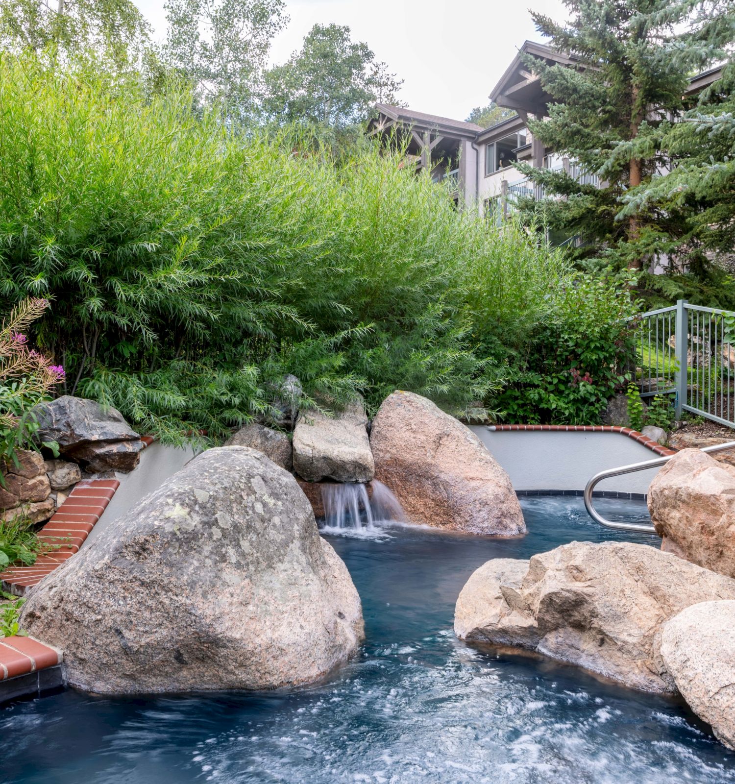 The image features a tranquil hot tub area surrounded by lush greenery, rocks, and a cozy backdrop of a building.