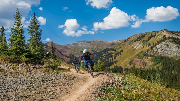 The image shows mountain bikers racing along a rocky trail surrounded by lush green trees and stunning mountain landscapes.