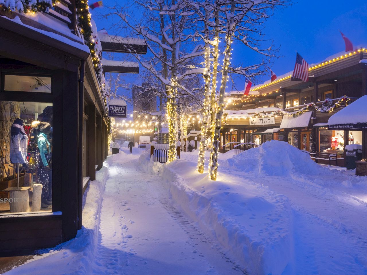 The image shows a snowy street decorated with lights and shops, creating a cozy winter scene in the evening.