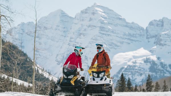 Two snowmobiles are parked in the snow, with two riders wearing helmets and colorful gear against a backdrop of snowy mountains.