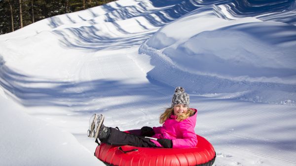 A smiling child is sitting on a red tube in snow, dressed in a pink jacket and gray hat, surrounded by winter scenery.