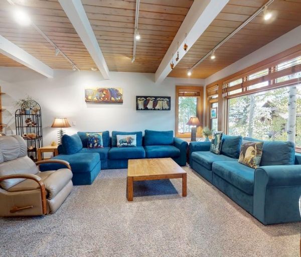 The image shows a cozy living room with teal sofas, a wooden coffee table, and large windows offering a scenic view outside.