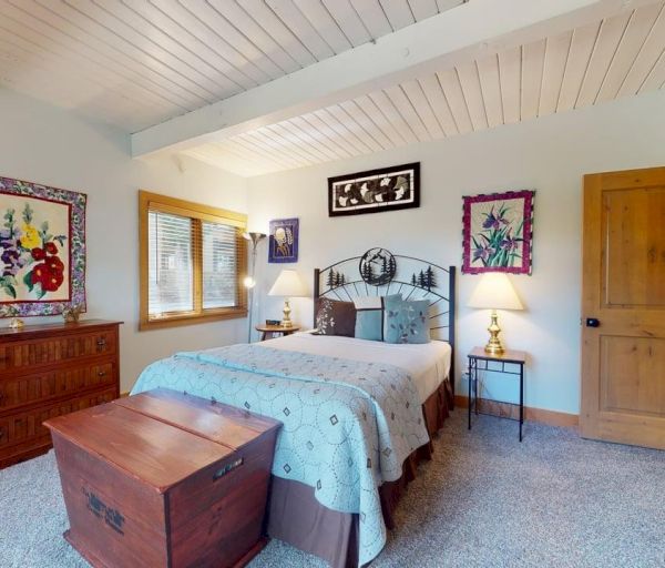 The image shows a cozy bedroom with a bed, decorative quilts, wooden furniture, lamps, and colorful artwork on the walls.