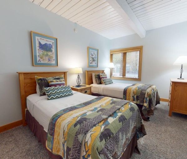 The image shows a cozy bedroom with two beds, colorful blankets, a nightstand, artwork on the wall, and a window for natural light.