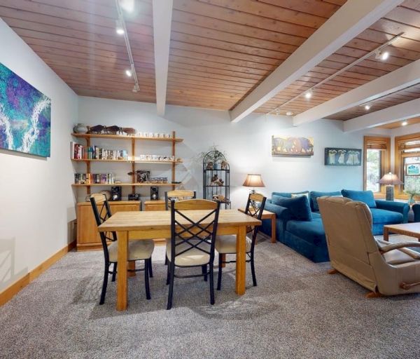 The image shows a cozy living area with a dining table, blue couches, wooden shelves, and artwork on the walls.
