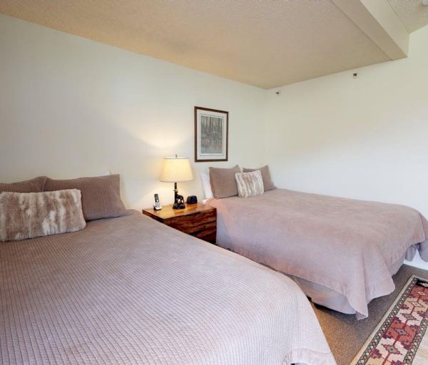 The image shows a cozy bedroom with two beds, a lamp, a nightstand, and a bathroom area visible in the background.