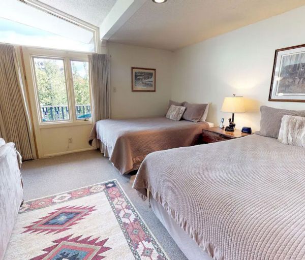 The image shows a cozy room with two beds, a sofa, and large windows letting in natural light. It&rsquo;s warm and inviting.