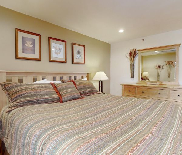 The image shows a cozy bedroom with a patterned bedspread, bedside lamps, framed artwork, and a wooden dresser with a mirror.