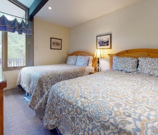 The image shows a cozy bedroom with two beds dressed in patterned bedding, a lamp, and large windows letting in natural light.