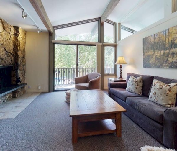 The image showcases a cozy living room with a stone fireplace, a sofa, a wooden coffee table, and large windows overlooking greenery.