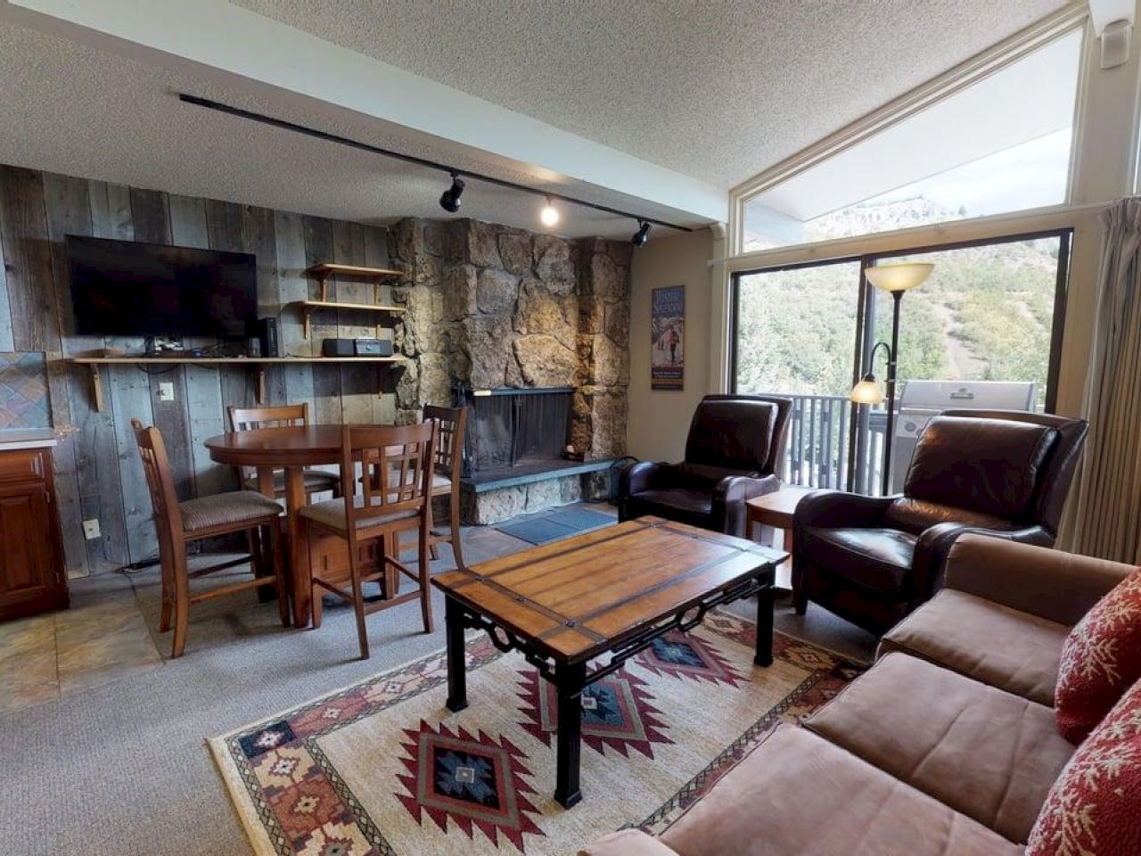 The image shows a cozy living area with a fireplace, comfortable seating, a dining table, and large windows revealing a scenic view.