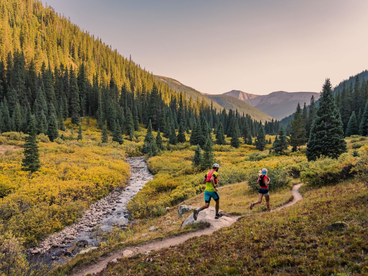 Two runners navigate a scenic trail through a vibrant valley surrounded by mountains and lush greenery. The sun is setting.
