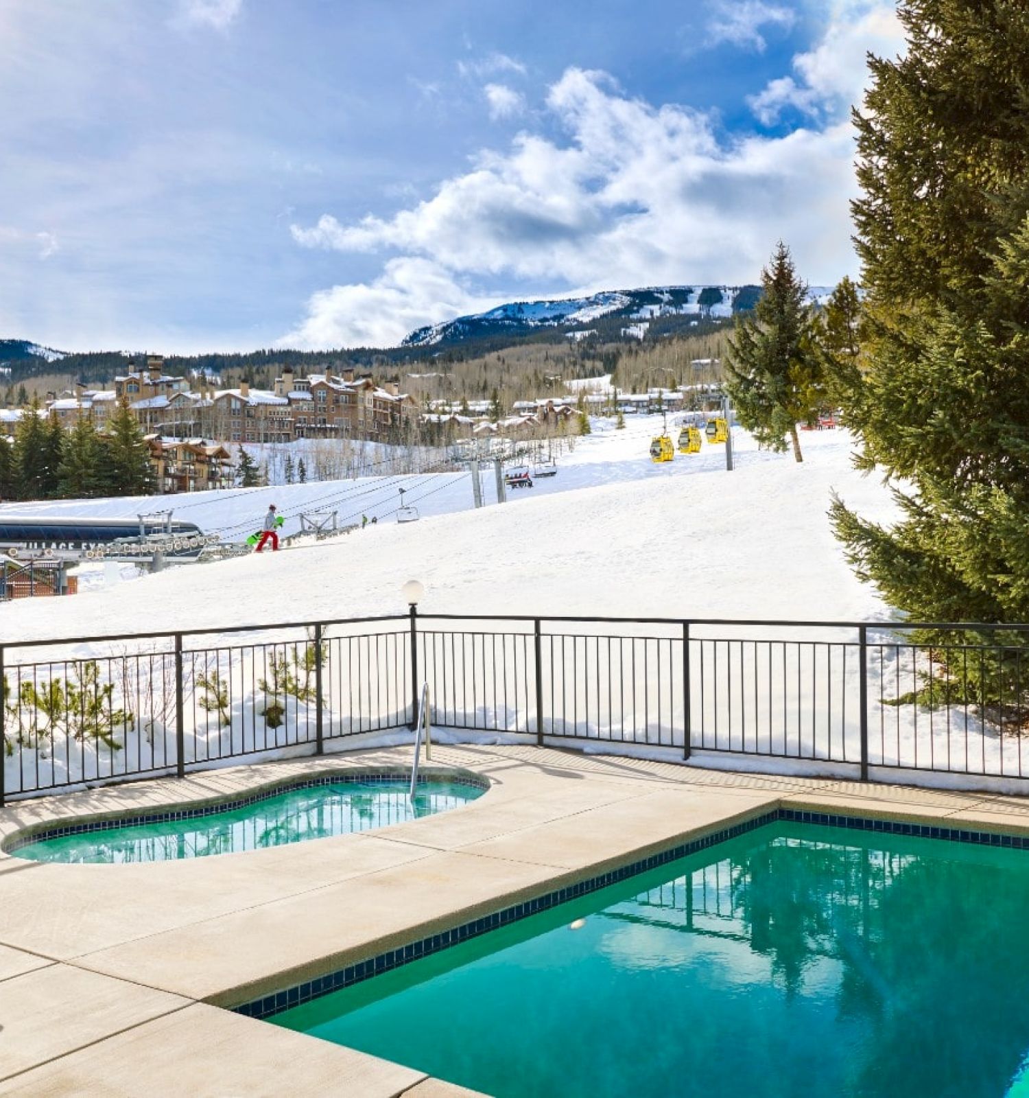 A sunny snowy slope view beyond a fenced pool area with two pools, a deck chair, and evergreen trees by a bright blue sky.