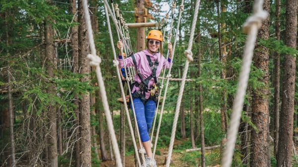 A person in a harness balances on a rope course among trees, wearing a helmet and smiling, enjoying an adventurous activity.