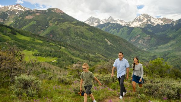 A family of three hiking on a hillside path in a lush mountain landscape, with snow-capped peaks in the distance.
