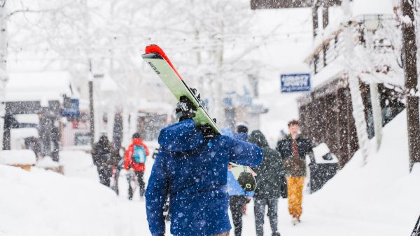 A person in a blue jacket carries skis while snow falls, surrounded by others in a snowy winter scene.