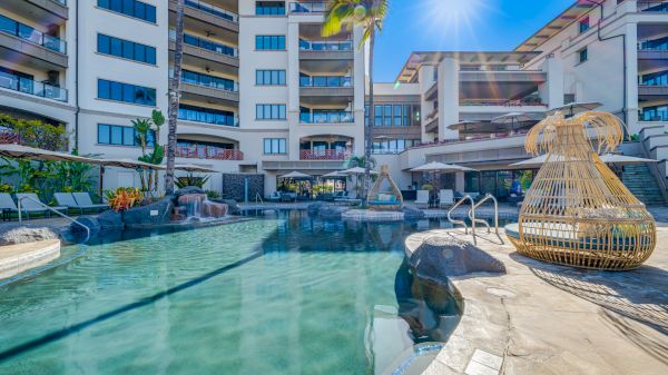 The image shows a luxurious pool area surrounded by a large multi-story building, with lounge chairs and a wicker cabana under a clear blue sky.