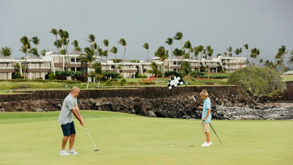 Two people are playing golf on a lush course, with palm trees and buildings in the background, under a cloudy sky.