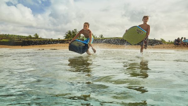 Two boys run through shallow water at the beach, each holding a bodyboard. A sunny day with palm trees and beachgoers in the background.