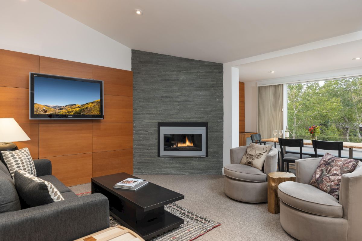 The image shows a modern living room with a TV, fireplace, cozy seating, and a dining area with views of greenery outside.