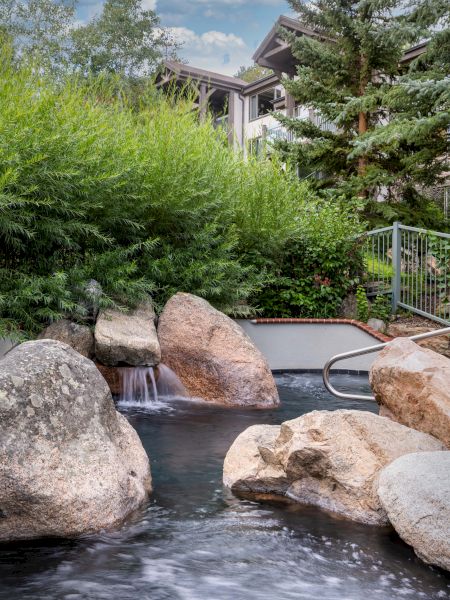 The image showcases a serene outdoor setting featuring a small waterfall, rocks, and lush greenery, alongside a cozy building.