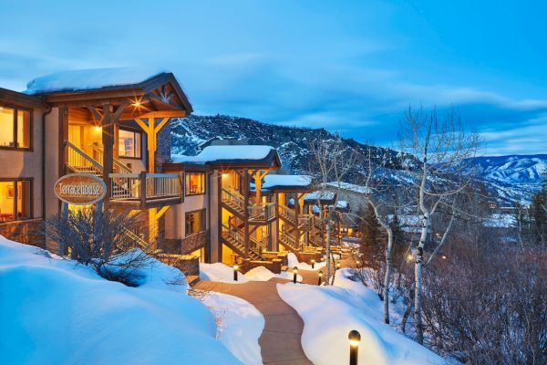 The image shows a modern mountain house surrounded by snow, with a scenic view and warm lights glowing from its windows.