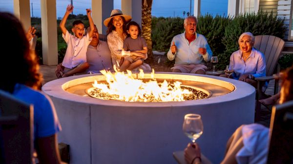 A group of people sits around a glowing fire pit on a patio, cheering and enjoying a social gathering at dusk.