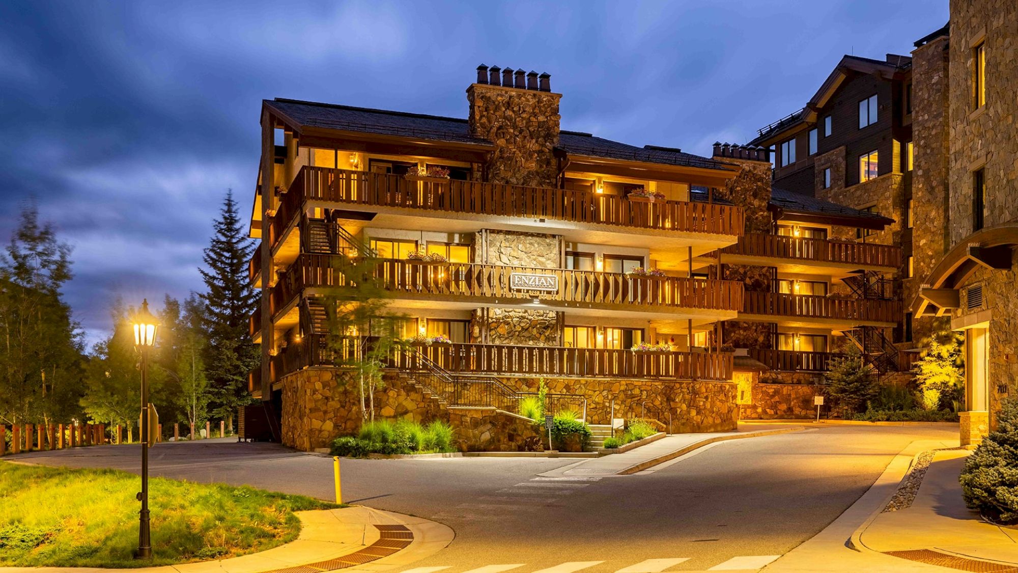 The image shows a cozy lodge with stone accents and wooden balconies, illuminated at dusk among trees and a winding road.