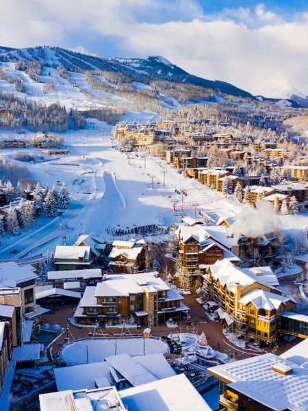 The image shows a picturesque snowy mountain resort with charming buildings, ski slopes, and trees blanketed in white snow.