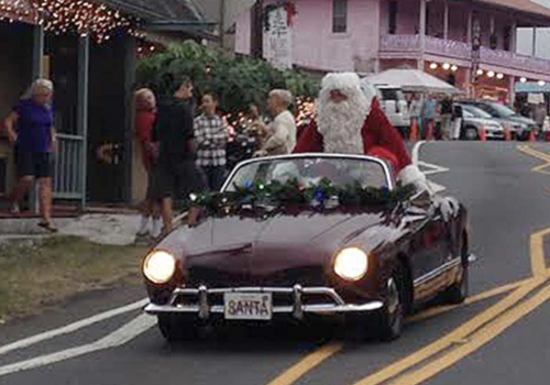 A classic maroon convertible driving Santa Claus down a street lined with holiday lights and onlookers, as a festive parade scene unfolds.